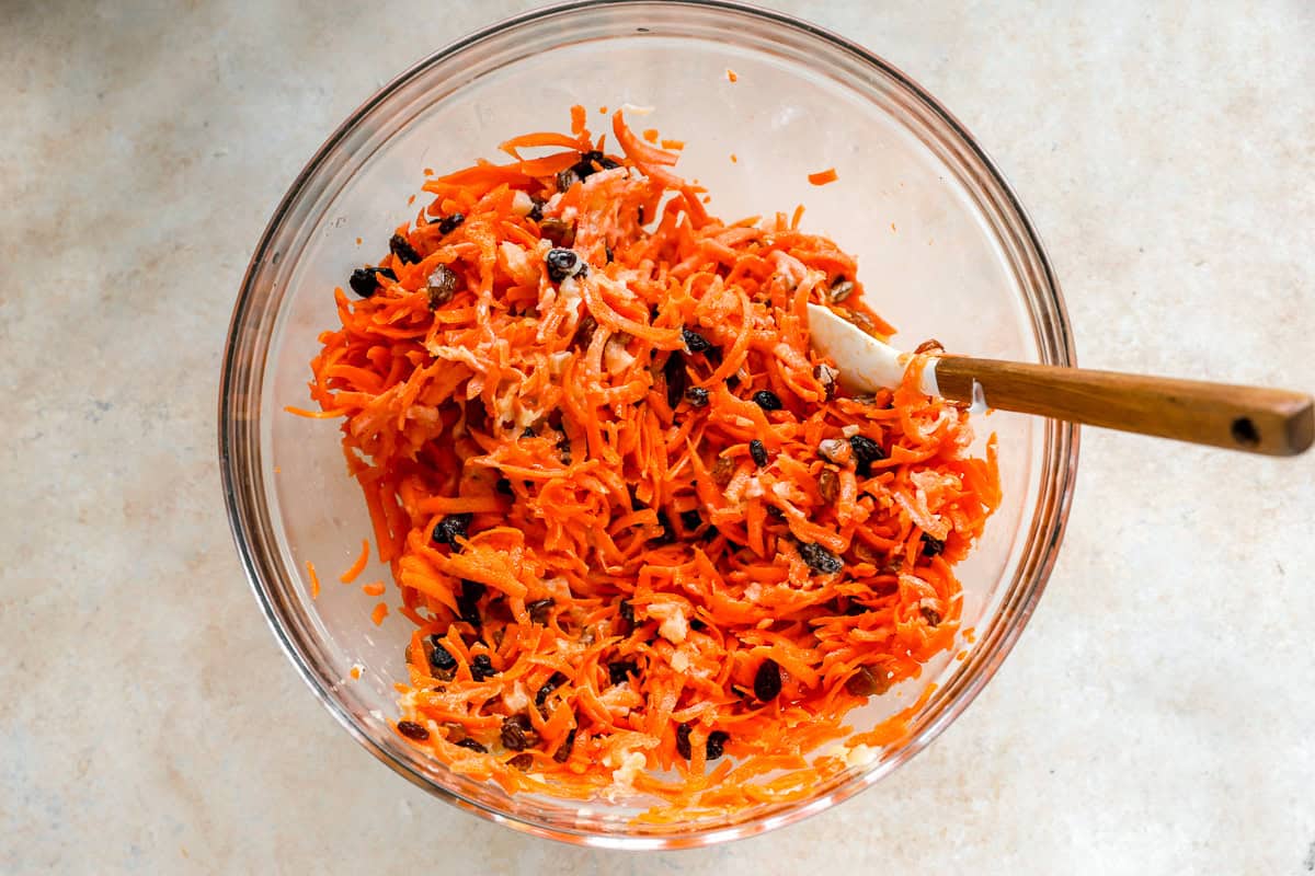 Glass mixing bowl of shredded carrot raisin salad being stirred with a spatula, showing visible raisins and pineapple pieces.