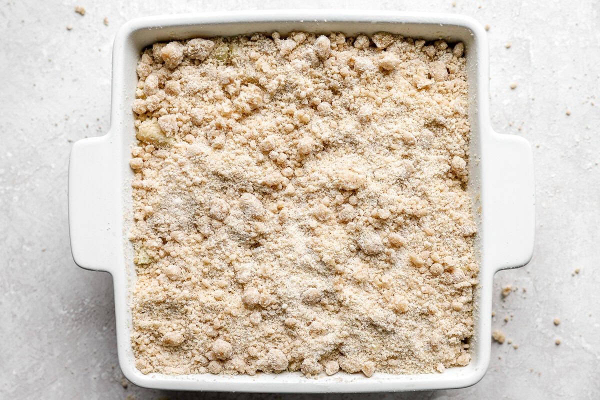White square baking dish with apples covered in an even layer of crumb topping before baking.