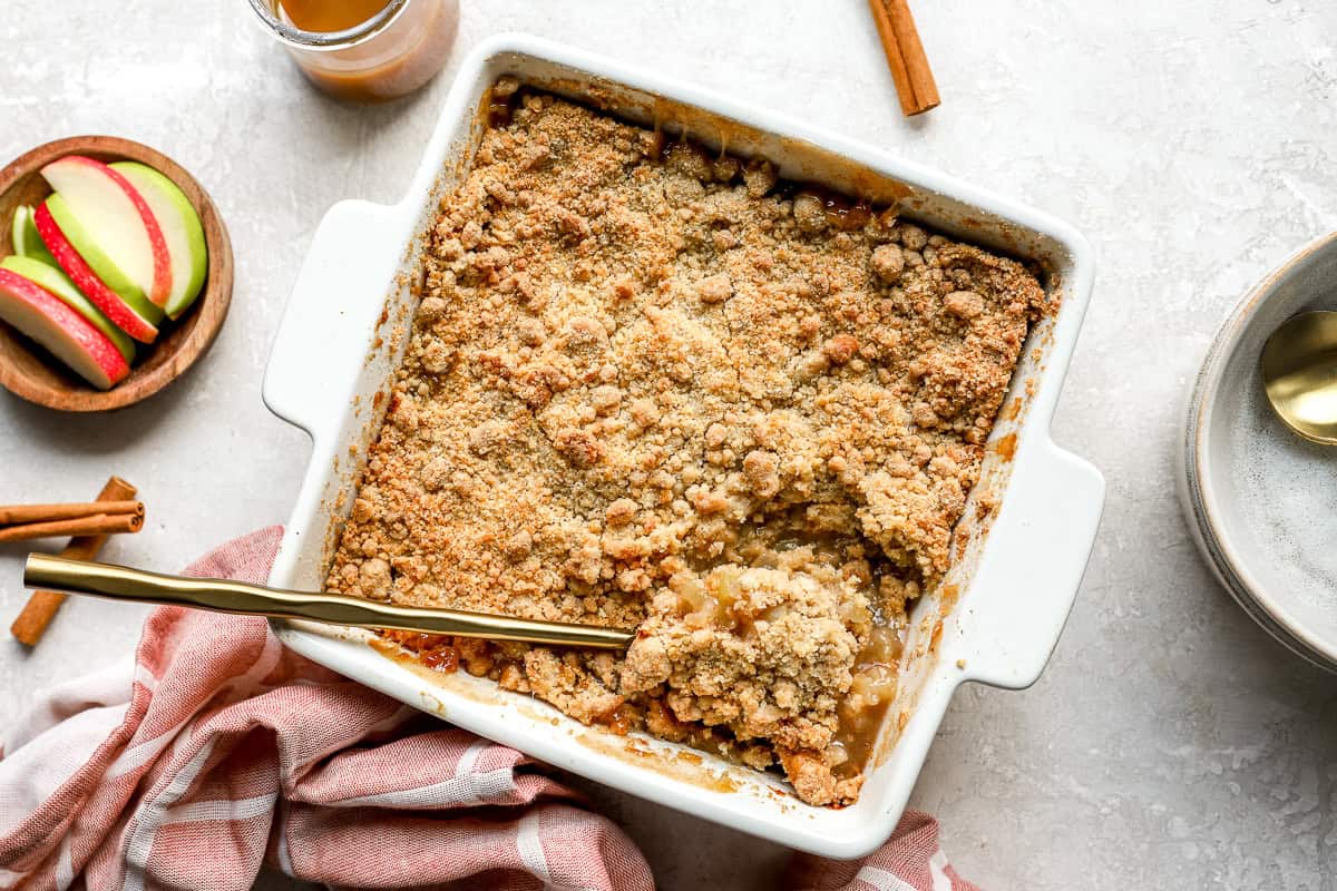 Freshly baked apple crumble in a white square baking dish with a golden brown topping, partially scooped to reveal filling.