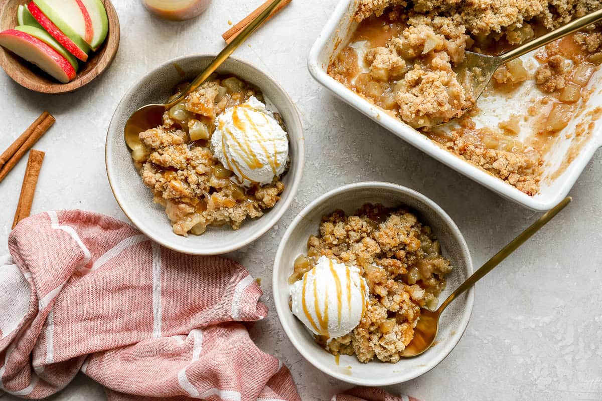 Two bowls of apple crumble with vanilla ice cream and caramel sauce next to a baking dish of crumble, gold spoons, and sliced apples on the side.