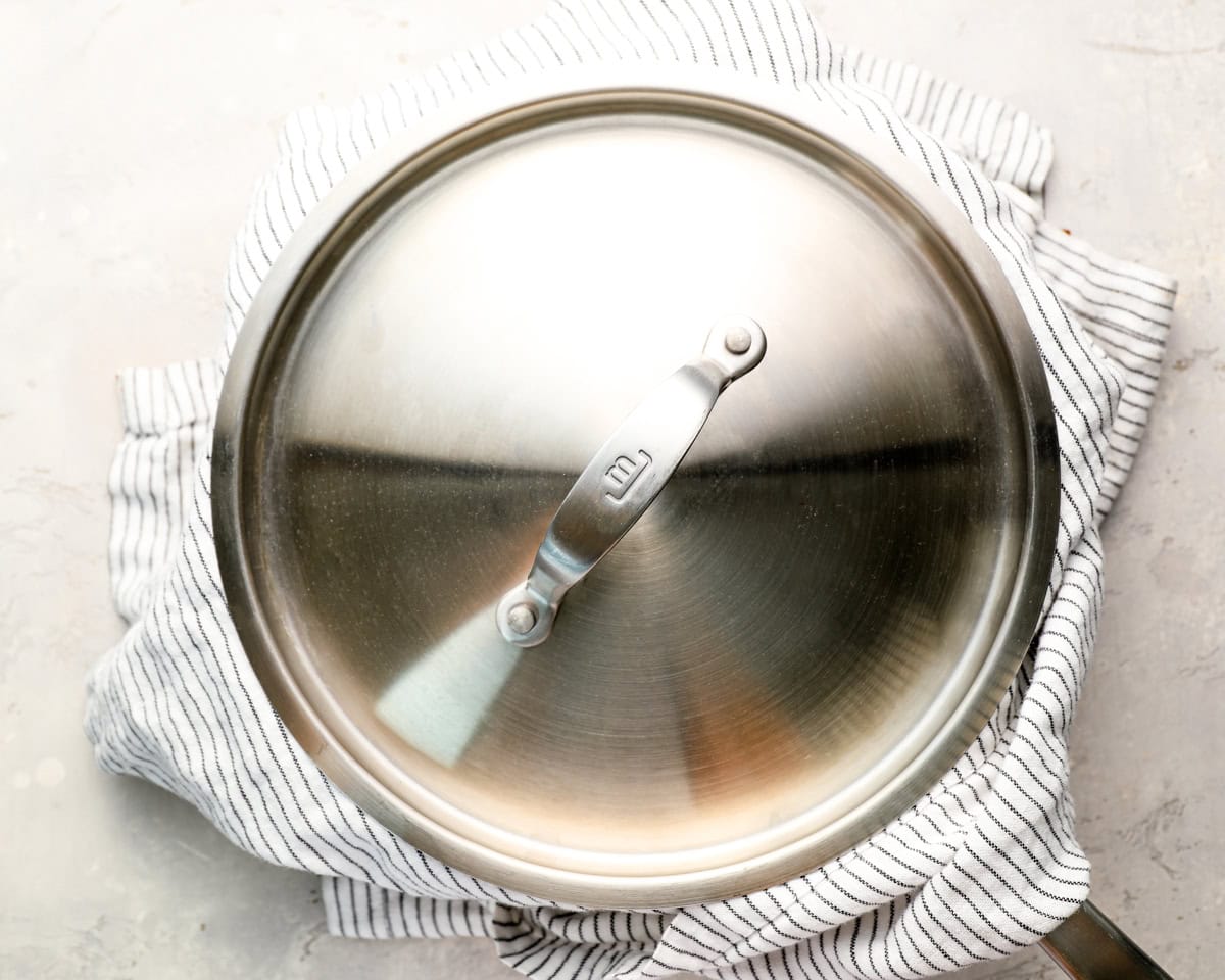 tainless steel skillet covered with a lid, resting on a white and black striped kitchen towel on a light countertop