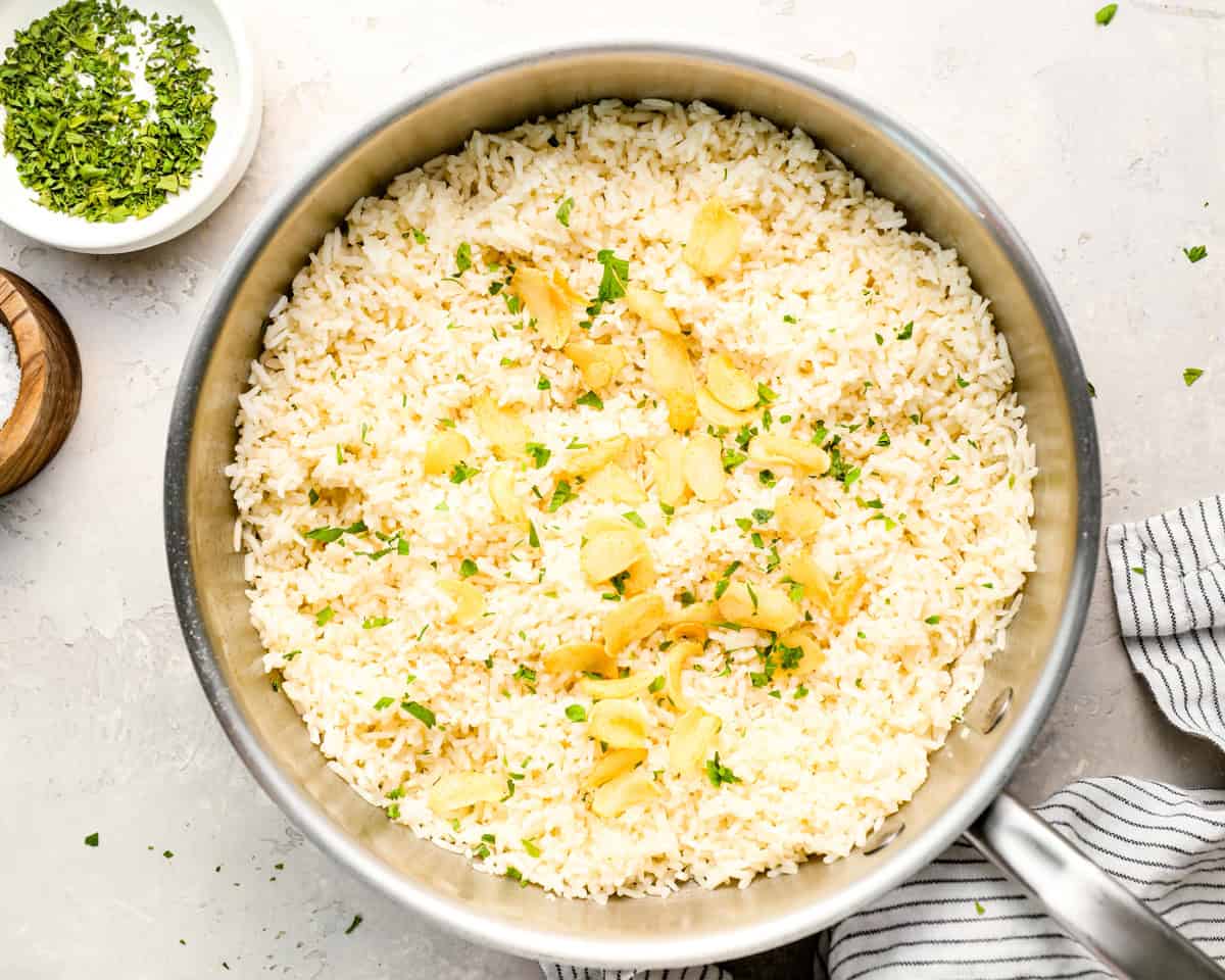 Cooked garlic rice in a stainless steel skillet, topped with golden garlic slices and chopped parsley, with small bowls of salt and herbs nearby on a light surface