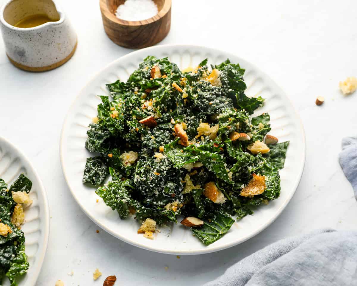 Plate of kale salad served with toppings scattered, with a small pitcher of dressing and salt in the background.