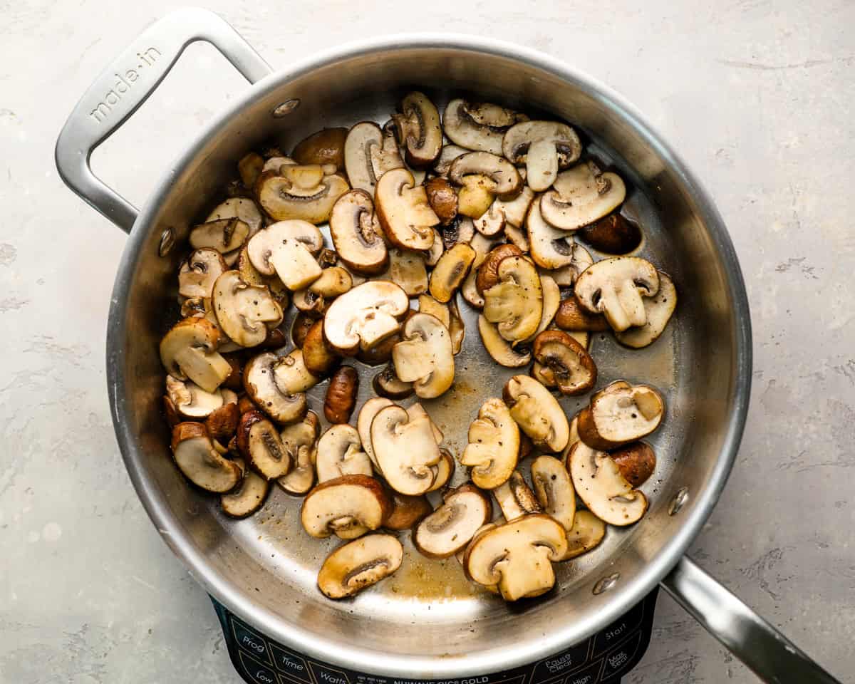 Raw sliced mushrooms cooking in a stainless steel skillet over medium heat, just beginning to release their moisture.