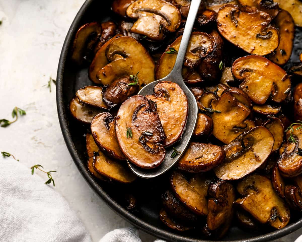 Close-up of a spoon lifting a slice of browned mushroom from a bowl of sautéed mushrooms, showing glossy texture and thyme leaves.