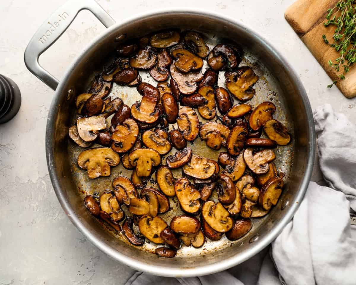Sautéed mushrooms fully browned and coated in glaze, filling a stainless steel skillet surrounded by thyme and a linen towel.
