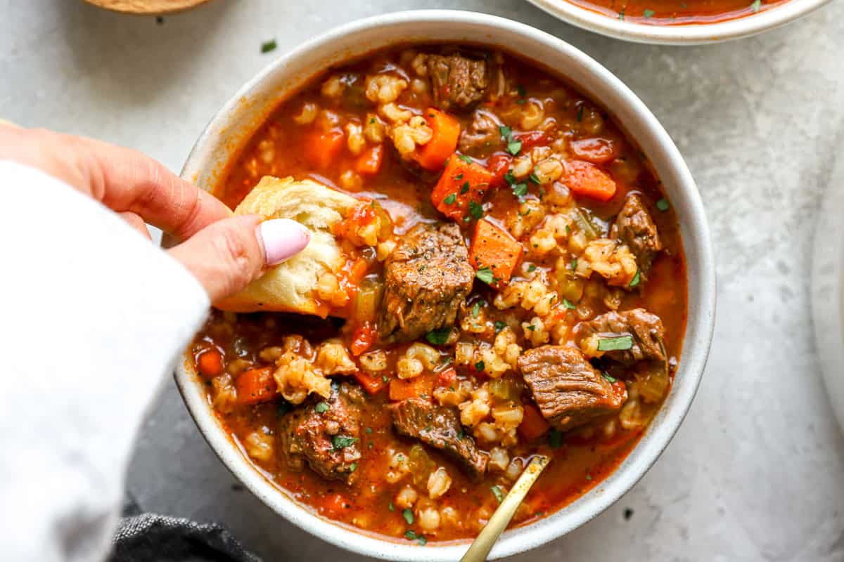 A hand dipping a piece of crusty bread into a bowl of rich beef barley soup with visible beef chunks and vegetables.