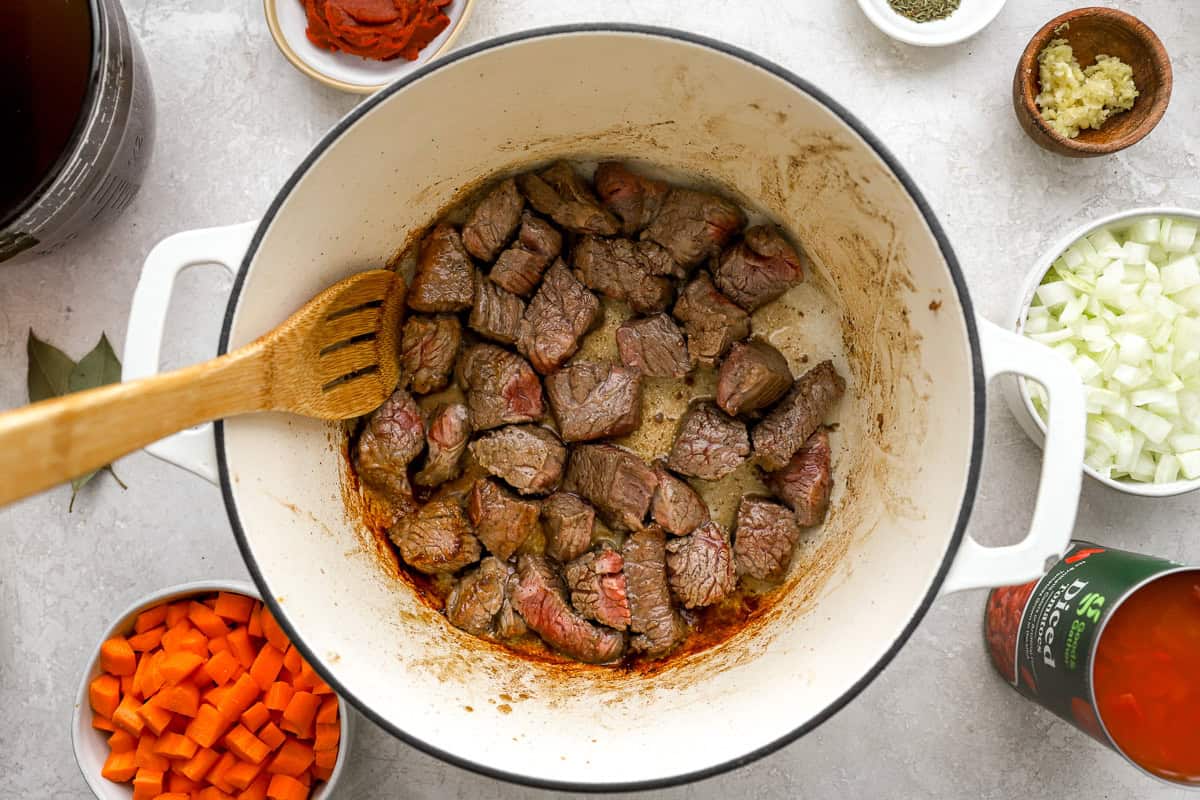 Cubed beef searing in a Dutch oven, surrounded by prep ingredients including tomato paste, garlic, onions, carrots, and diced tomatoes.