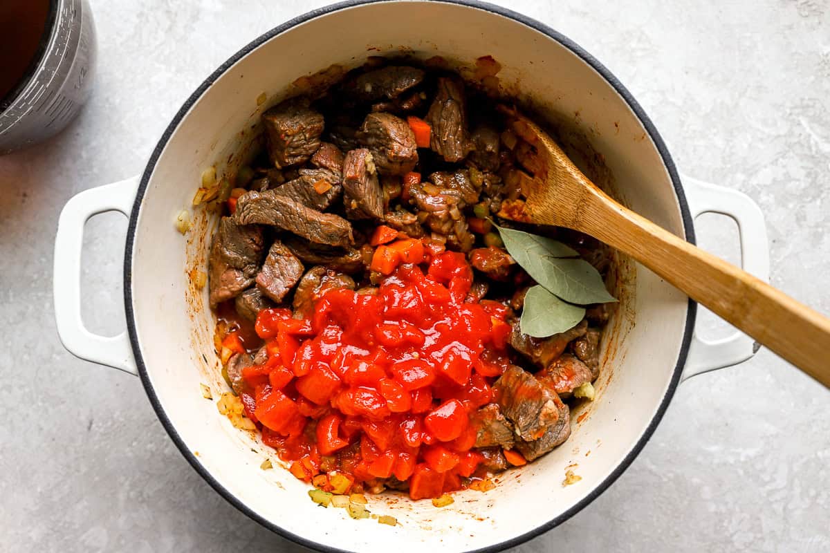 Diced tomatoes and bay leaves added to browned beef and sautéed vegetables in a Dutch oven before simmering.
