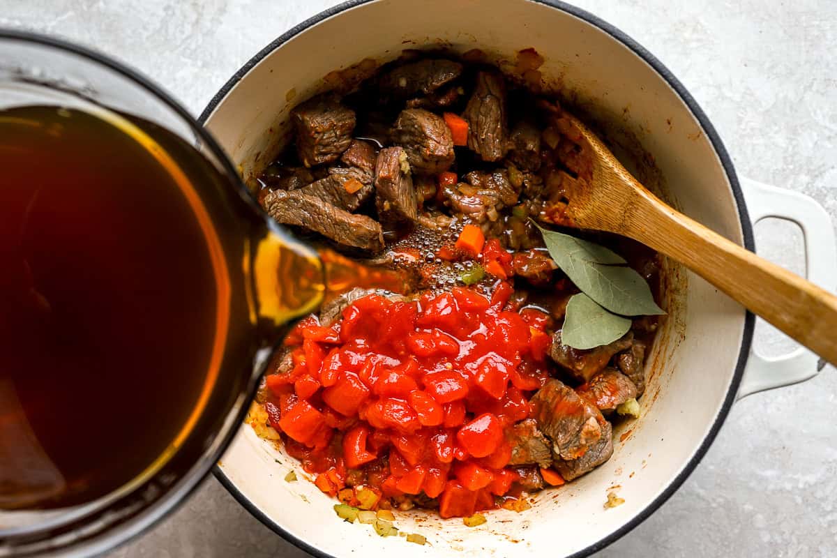 Beef broth being poured into the Dutch oven over the mixture of beef, tomatoes, and vegetables.