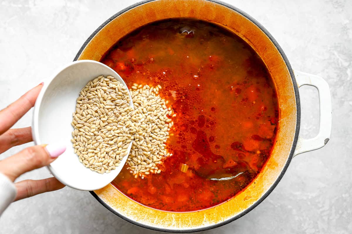 A bowl of pearled barley being added to the simmering soup base in a Dutch oven.