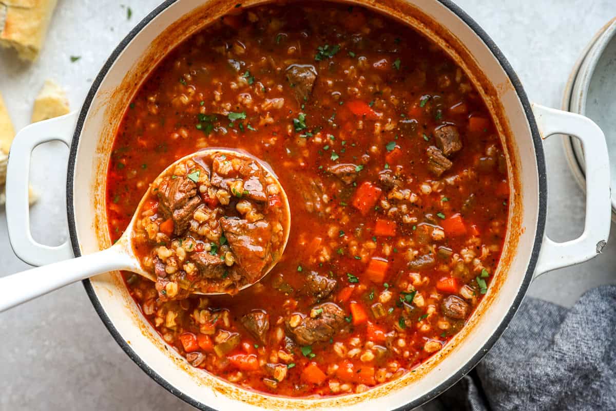 A ladle full of beef barley soup lifted from a Dutch oven, showing chunks of beef, barley, carrots, and herbs.
