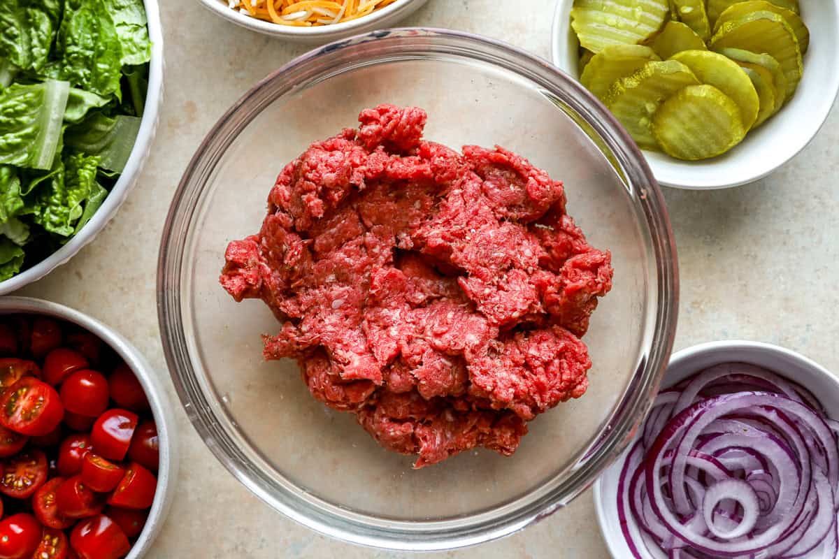 Glass mixing bowl filled with raw ground beef surrounded by bowls of shredded cheese, pickles, sliced red onions, grape tomatoes, and romaine lettuce.