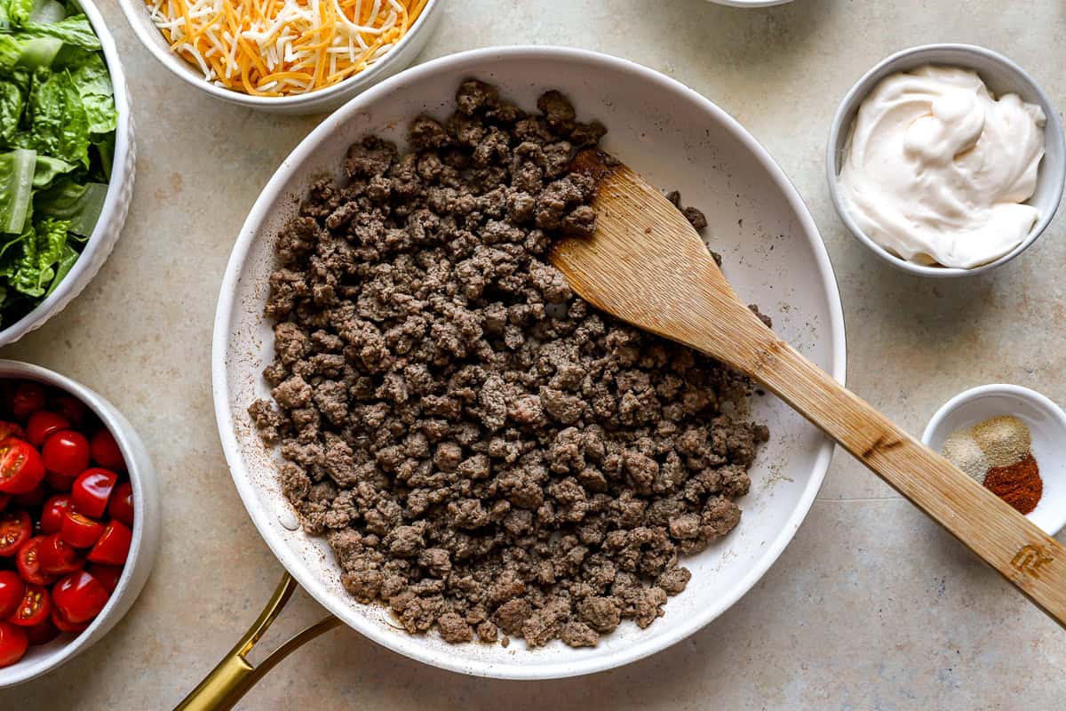 Cooked ground beef in a white skillet with a wooden spoon, surrounded by bowls of shredded cheese, mayonnaise, grape tomatoes, and seasoning spices.