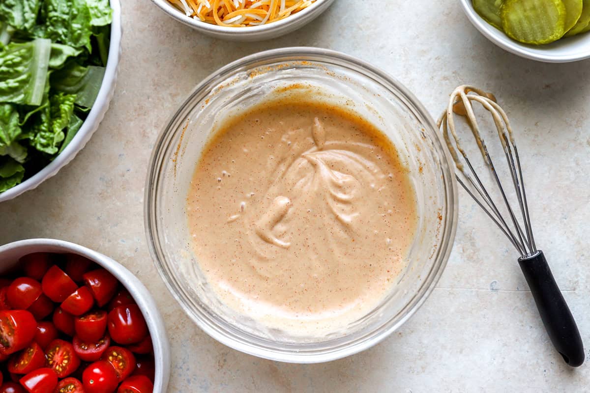 Creamy burger sauce whisked in a glass bowl, surrounded by bowls of shredded cheese, lettuce, grape tomatoes, and pickle slices.