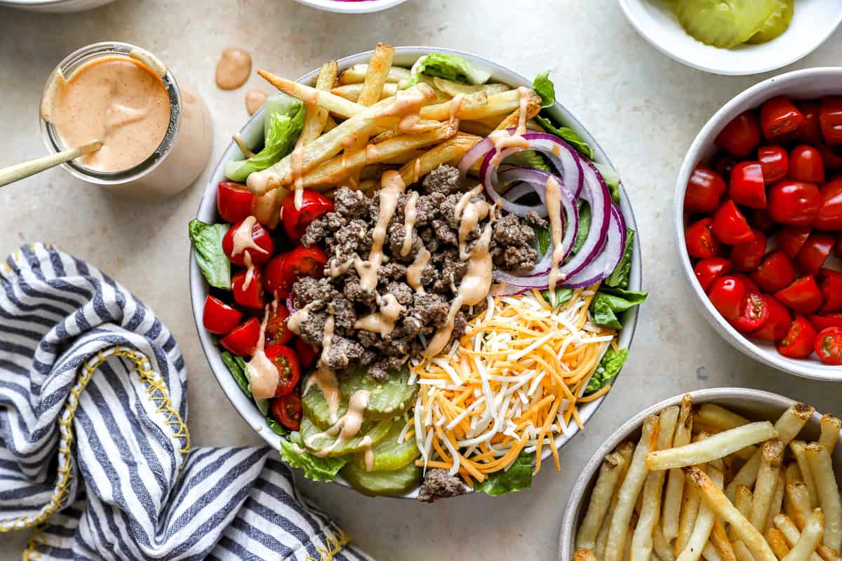Burger bowl topped with ground beef, fries, cheese, tomatoes, onions, pickles, and sauce, next to a jar of burger sauce, striped kitchen towel, and bowls of toppings.