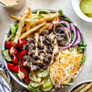 Overhead view of a burger bowl filled with ground beef, shredded cheddar cheese, lettuce, grape tomatoes, red onion slices, dill pickles, French fries, and drizzled burger sauce.