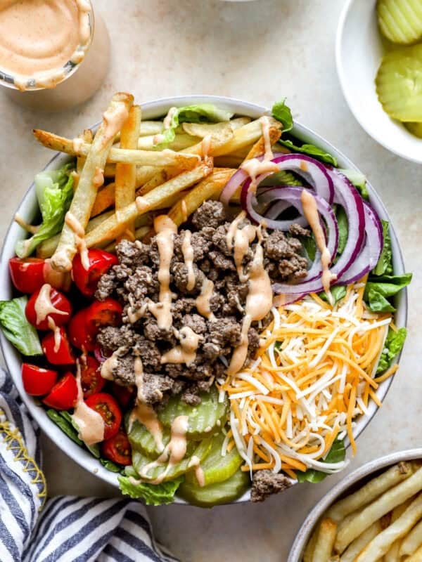 Overhead view of a burger bowl filled with ground beef, shredded cheddar cheese, lettuce, grape tomatoes, red onion slices, dill pickles, French fries, and drizzled burger sauce.