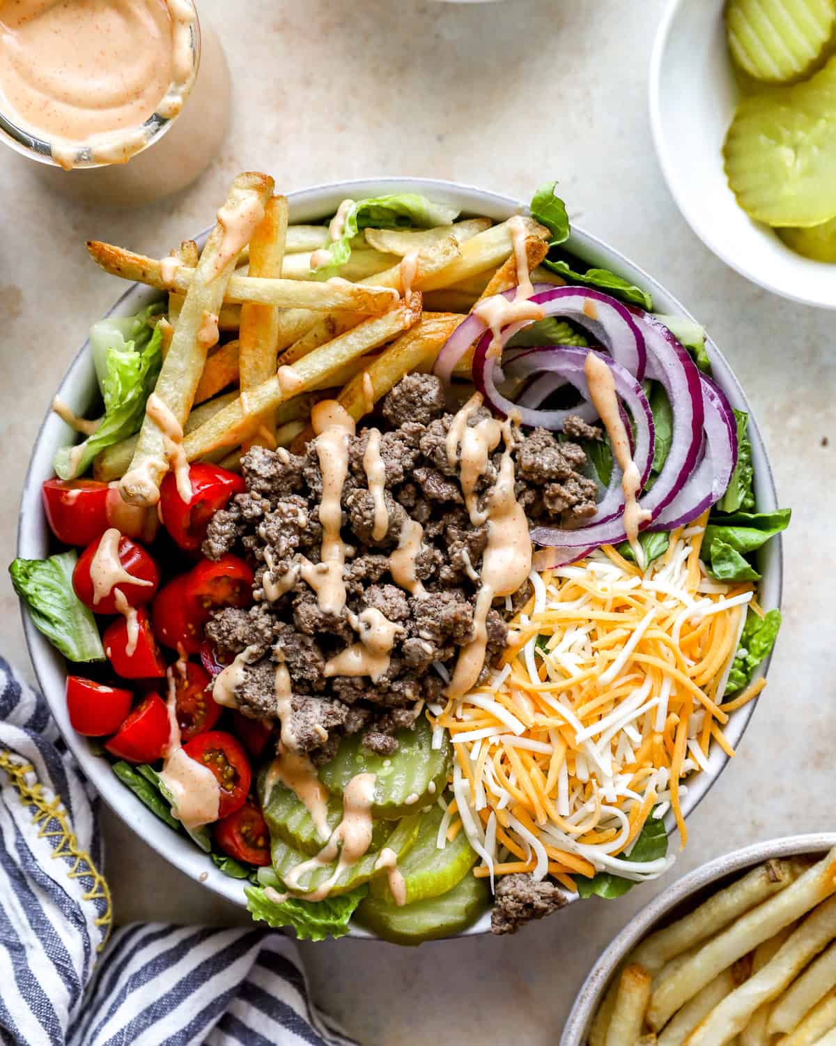 Overhead view of a burger bowl filled with ground beef, shredded cheddar cheese, lettuce, grape tomatoes, red onion slices, dill pickles, French fries, and drizzled burger sauce.