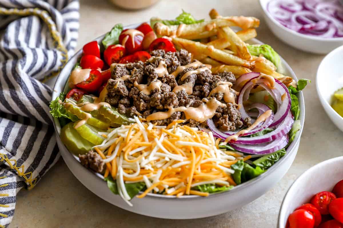 Side view of a burger bowl showing layers of lettuce, ground beef, shredded cheese, pickles, tomatoes, red onion, and crispy fries drizzled with sauce.