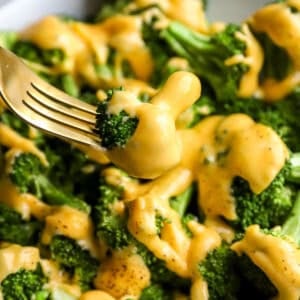 Close-up of a fork holding a broccoli floret coated in creamy cheddar cheese sauce over a plate of broccoli.