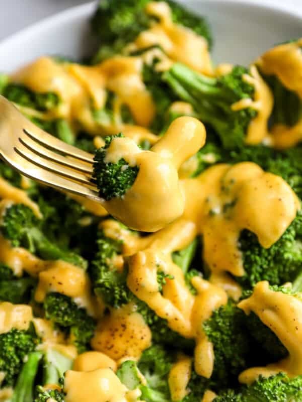 Close-up of a fork holding a broccoli floret coated in creamy cheddar cheese sauce over a plate of broccoli.