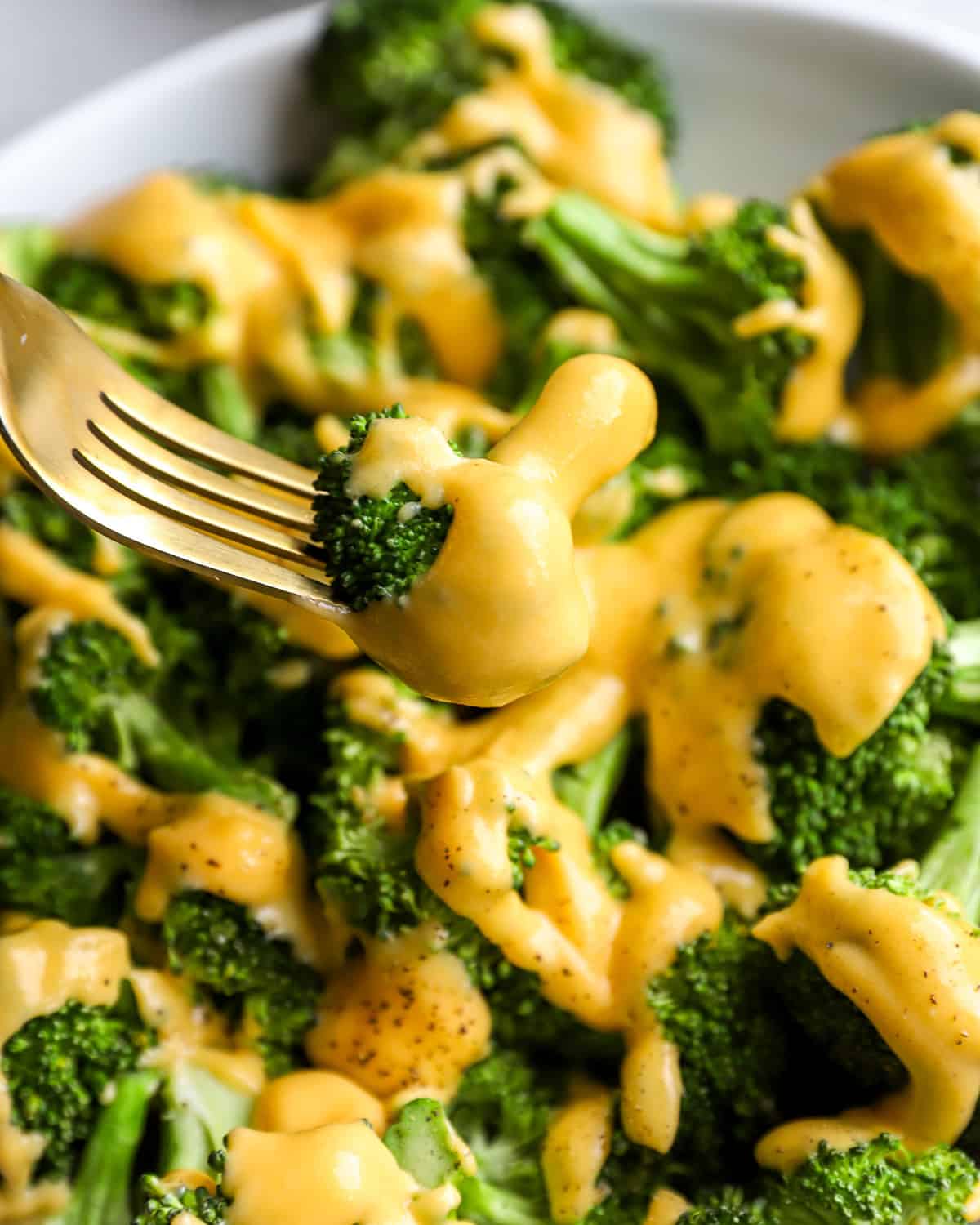 Close-up of a fork holding a broccoli floret coated in creamy cheddar cheese sauce over a plate of broccoli.