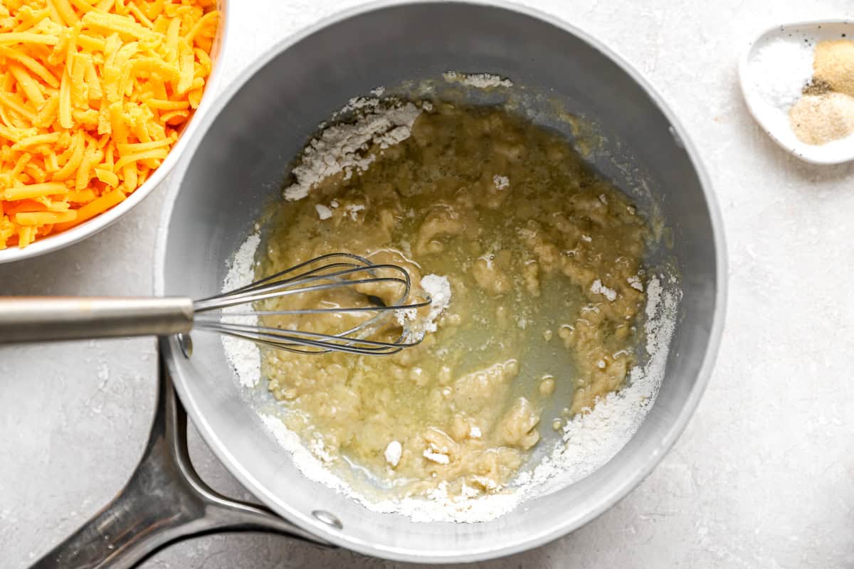 Melted butter and flour whisked together in a saucepan to form a roux, with shredded cheddar cheese in a bowl nearby.
