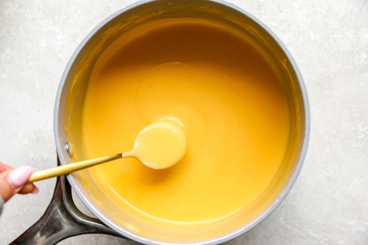 Smooth, velvety cheddar cheese sauce in a saucepan with a spoon resting inside.