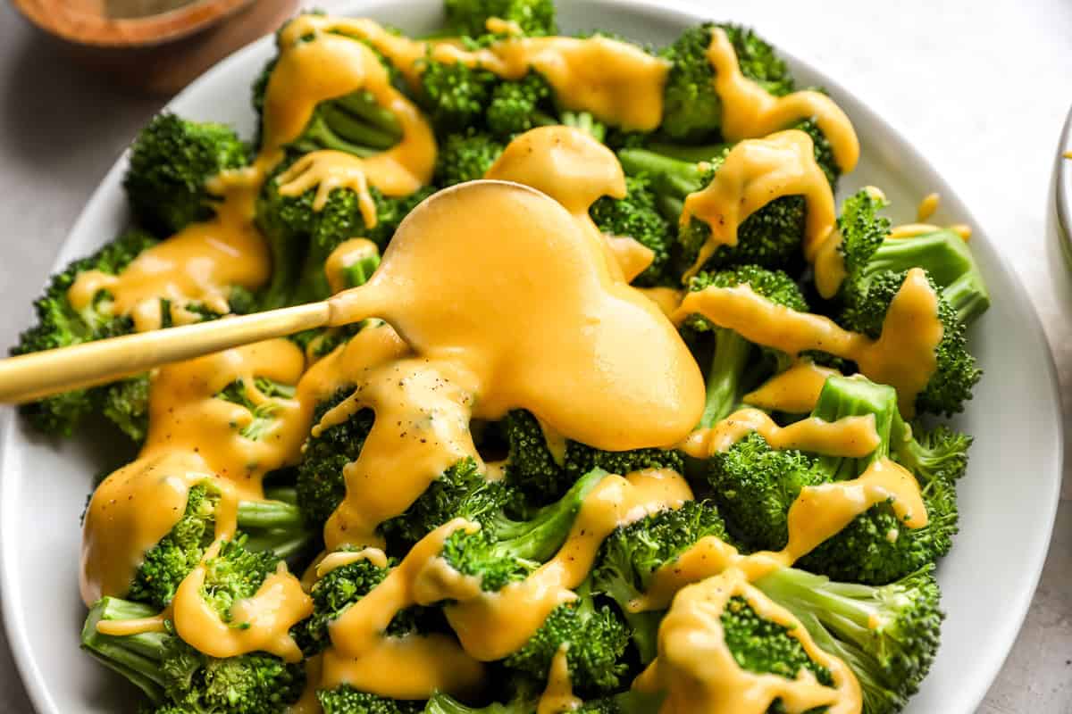 Spoon pouring creamy cheddar cheese sauce over a plate of steamed broccoli.