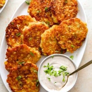 Golden brown corn fritters on a white platter topped with green onions, served with a bowl of sour cream dip.