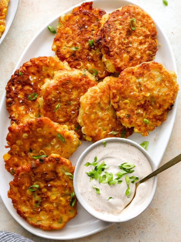 Golden brown corn fritters on a white platter topped with green onions, served with a bowl of sour cream dip.
