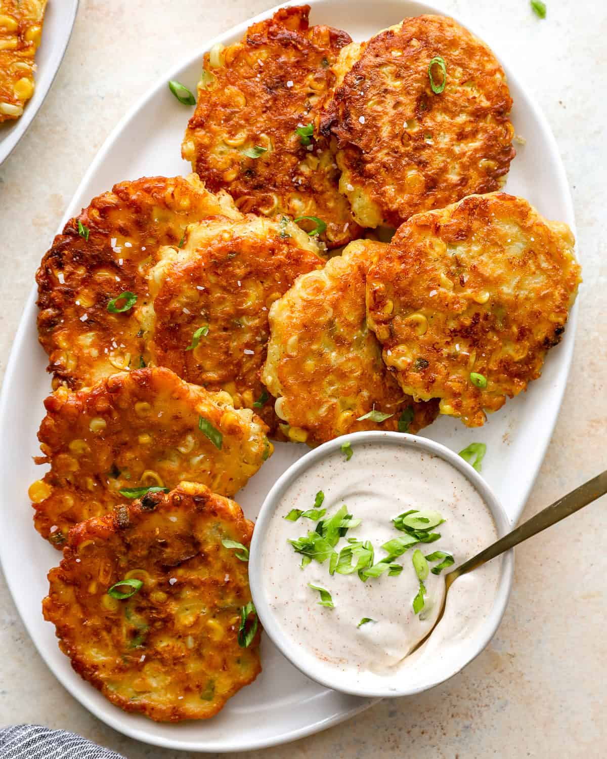 Golden brown corn fritters on a white platter topped with green onions, served with a bowl of sour cream dip.