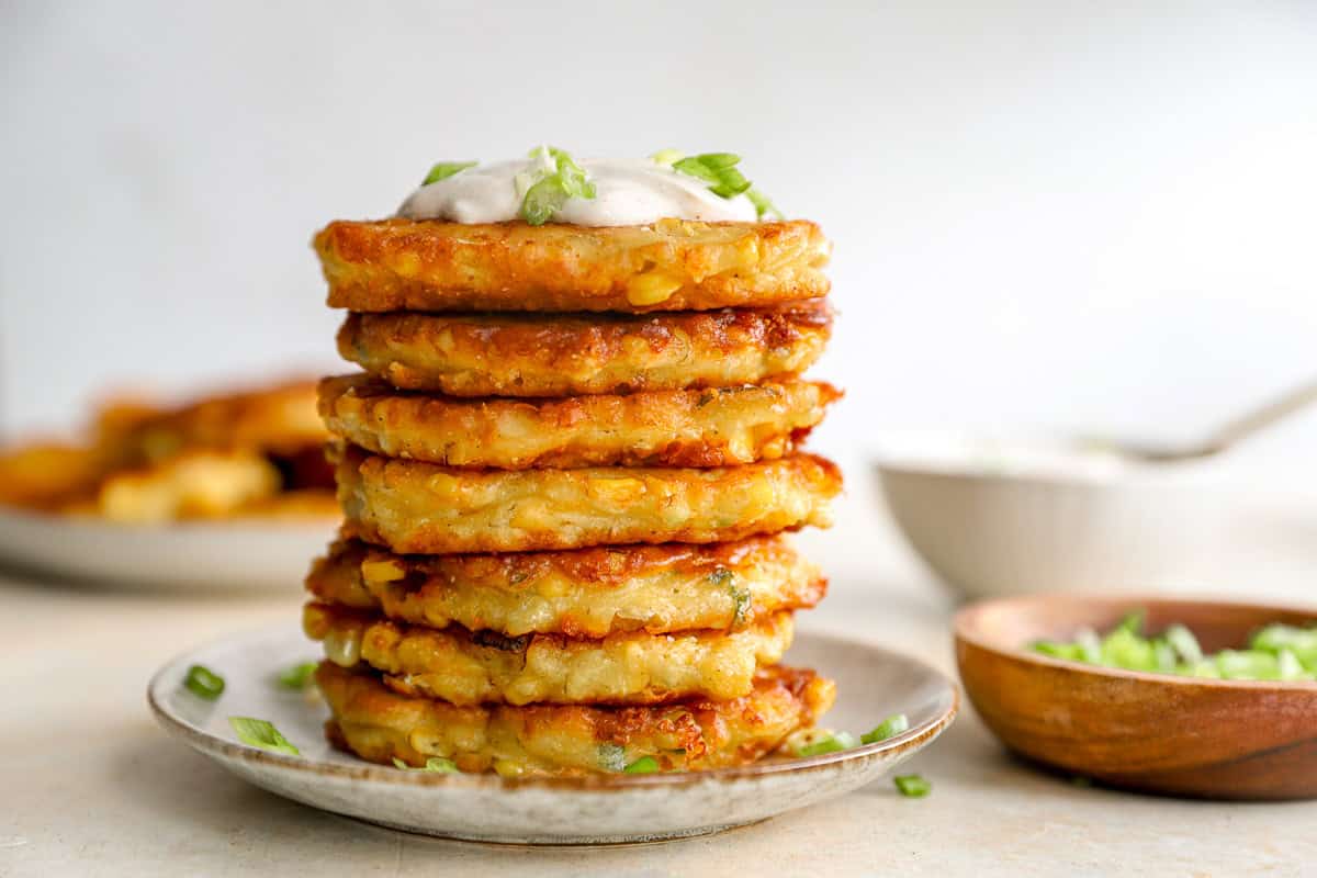 A tall stack of corn fritters on a plate topped with sour cream and green onions.