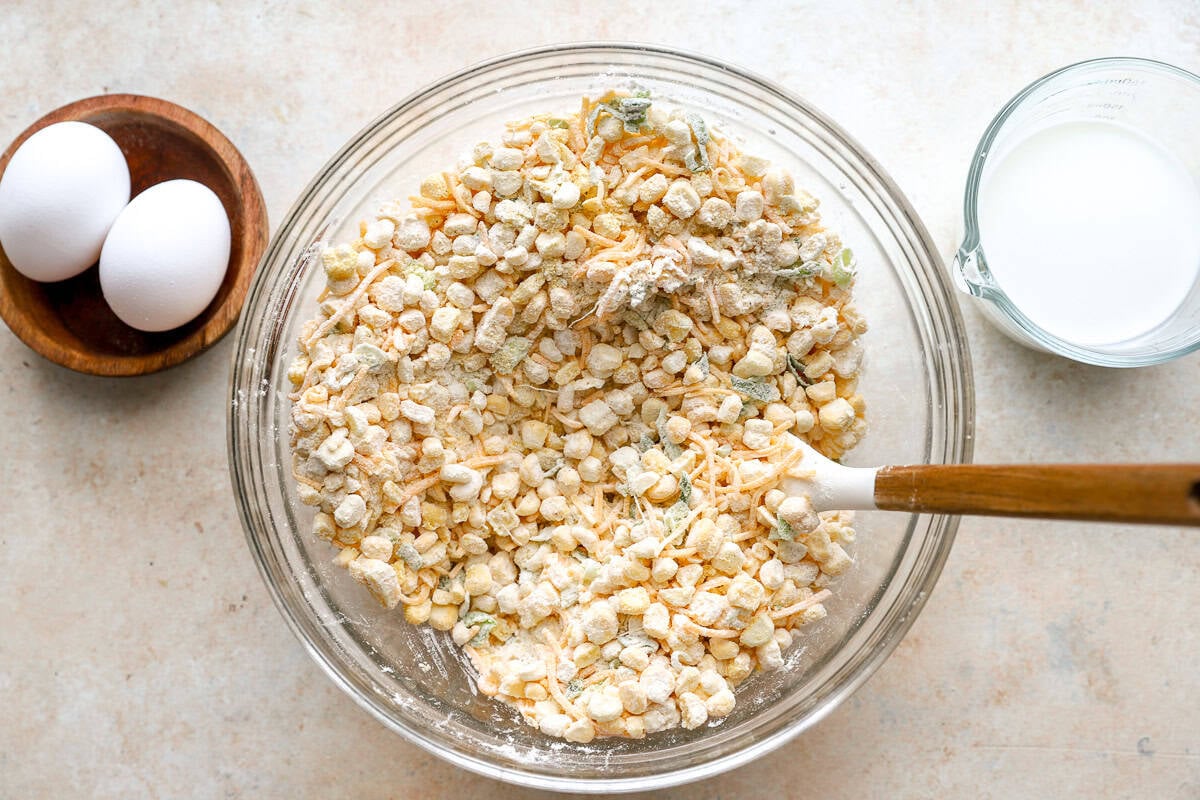 Mixing bowl filled with corn, shredded cheese, flour, and green onions before adding the wet ingredients, with eggs and milk on the side.