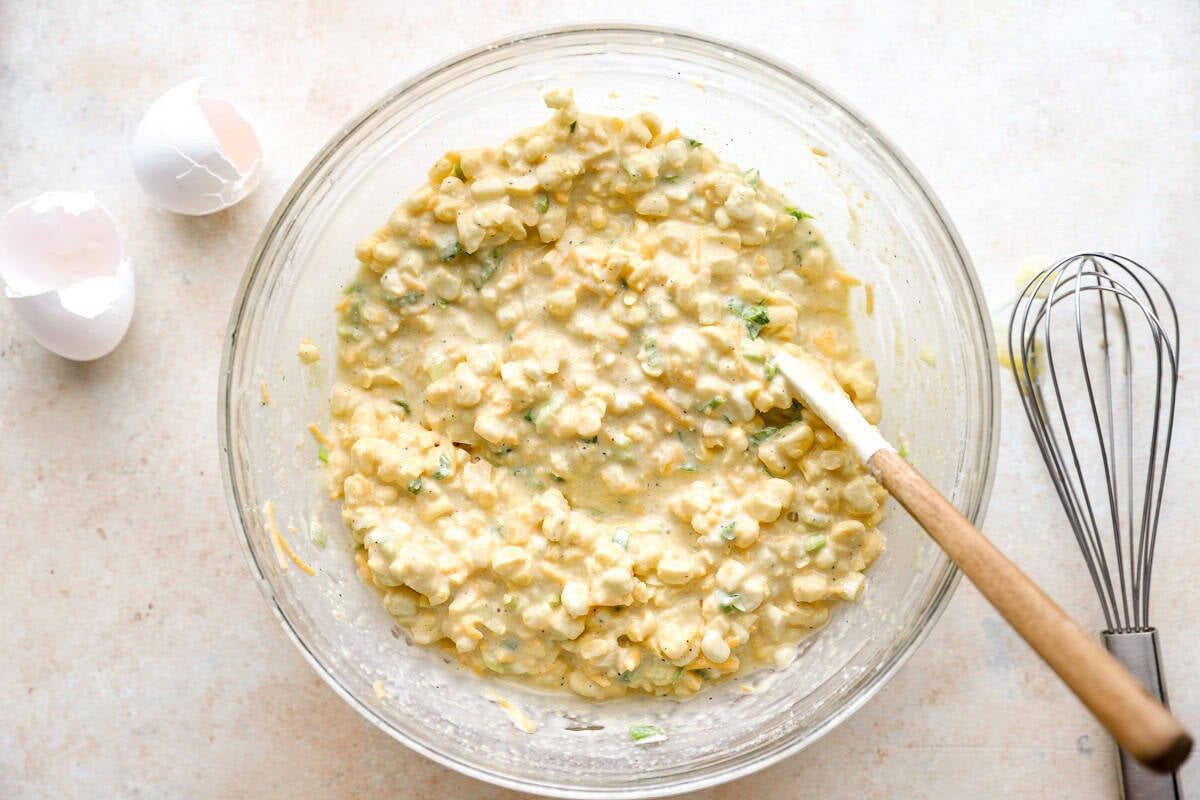 Corn fritter batter with corn, cheese, and green onions stirred together in a glass bowl with a whisk nearby.