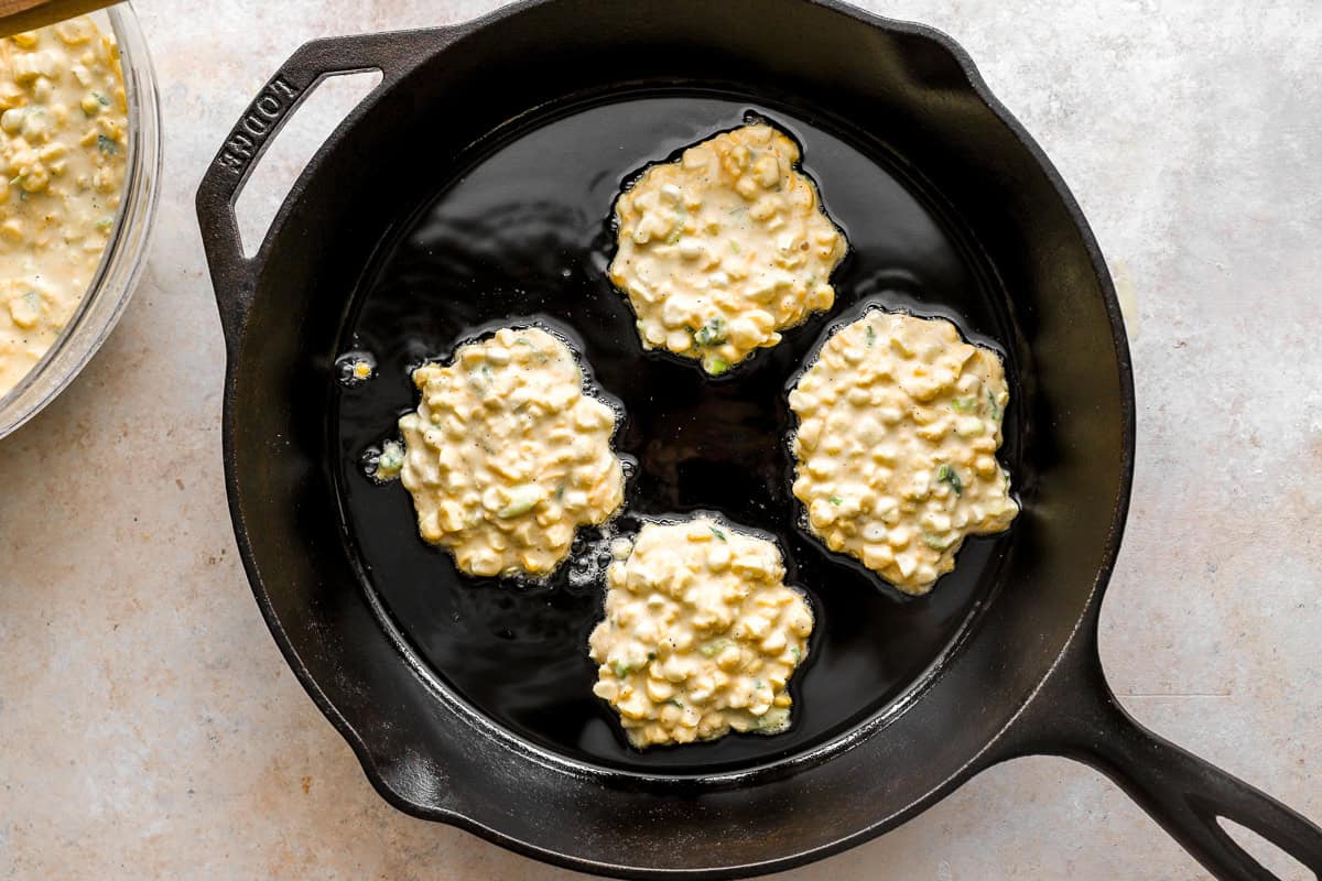 Four scoops of corn fritter batter frying in oil in a black cast iron skillet.