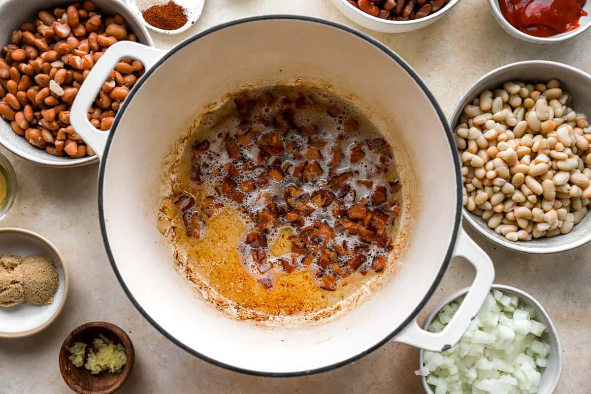 Large white Dutch oven with diced bacon sizzling in rendered fat, surrounded by bowls of beans, onion, garlic, brown sugar, spices, and ketchup.
