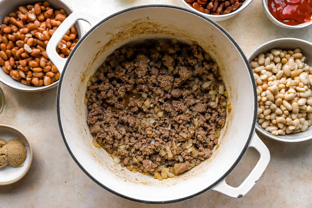White Dutch oven with browned ground beef and diced onions, with surrounding bowls of beans, seasonings, and sauce ingredients.