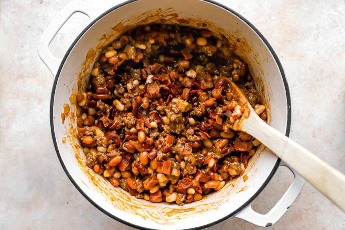 White Dutch oven filled with cooked ground beef, beans, and crisp bacon pieces, being stirred with a wooden spoon.