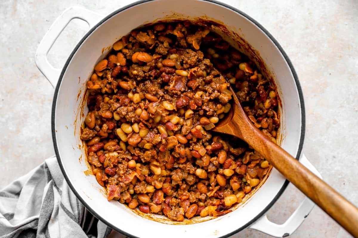 Overhead view of cowboy beans in a Dutch oven, showing a thick, saucy mix of beans, beef, and bacon with a wooden spoon resting in the pot.