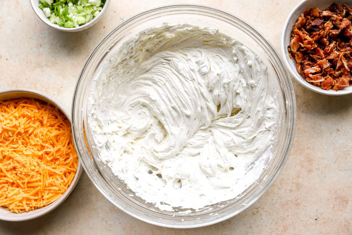 Cream cheese, sour cream, and ranch seasoning whipped together in a mixing bowl with bowls of cheddar, bacon, and green onions nearby.