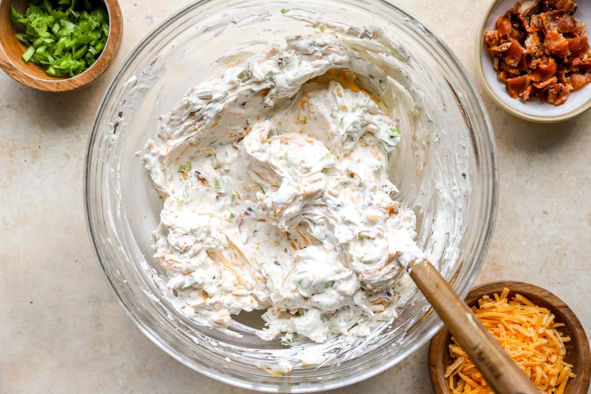 Crack dip being mixed together with a spatula, showing the creamy texture with bacon, cheese, and green onions throughout.