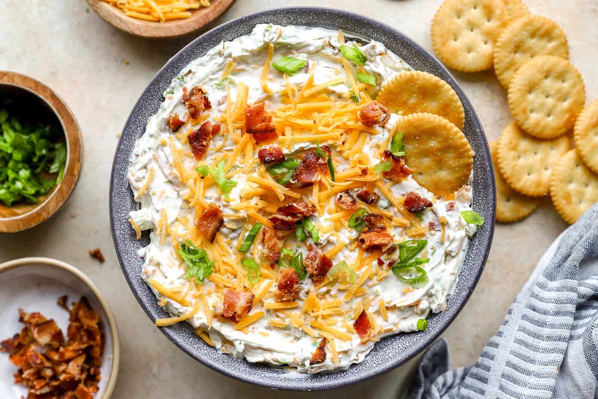 Overhead view of finished crack dip in a bowl, topped with bacon, cheddar, and green onions, served with round crackers.
