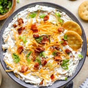Finished crack dip topped with shredded cheddar, bacon, and green onions in a bowl with crackers on the side.