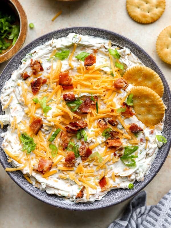 Finished crack dip topped with shredded cheddar, bacon, and green onions in a bowl with crackers on the side.