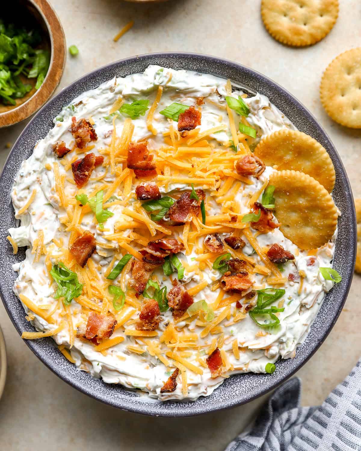 Finished crack dip topped with shredded cheddar, bacon, and green onions in a bowl with crackers on the side.