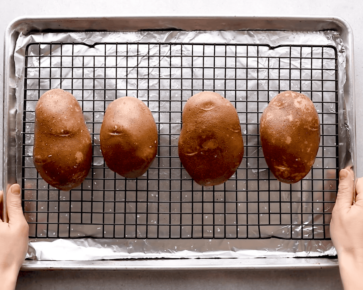 Four Russet potatoes rubbed with oil and seasoned, sitting on a wire rack over a foil-lined baking sheet before baking.