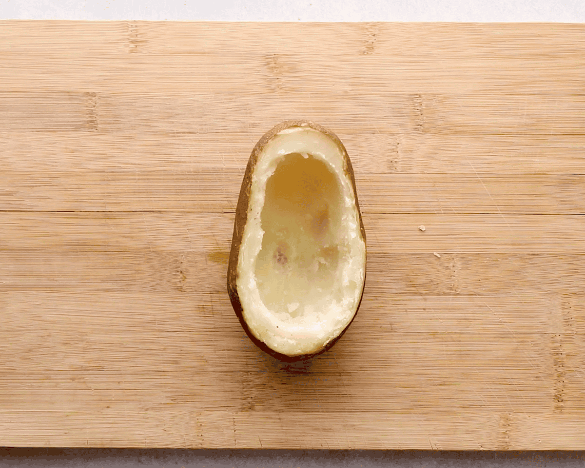 A halved potato with the inside scooped out, showing a clean potato shell on a wooden cutting board.