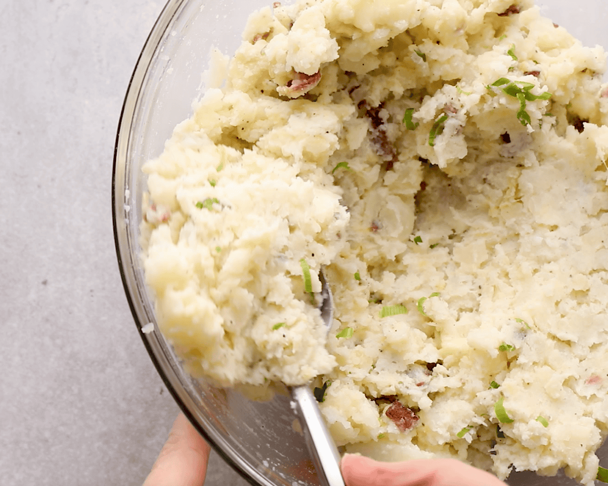Mashed potato mixture with bacon and scallions in a glass mixing bowl, being stirred with a spoon.